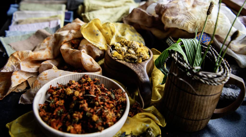 dried flowers in wooden bowls sit amongst scrunched piles of naturally dyed fabrics. A wooden cup contains plantain leaves.